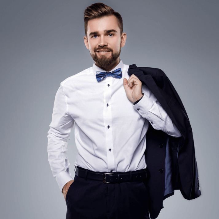 Mann im Anzug mit Fliege, hält seinen Blazer und lächelt vor grauem Hintergrund.