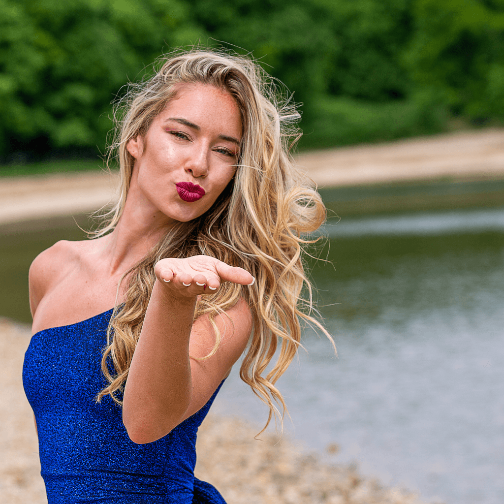 Frau mit blonden Haaren, die in einem blauen Kleid am Wasser steht und Küsse in die Kamera wirft.